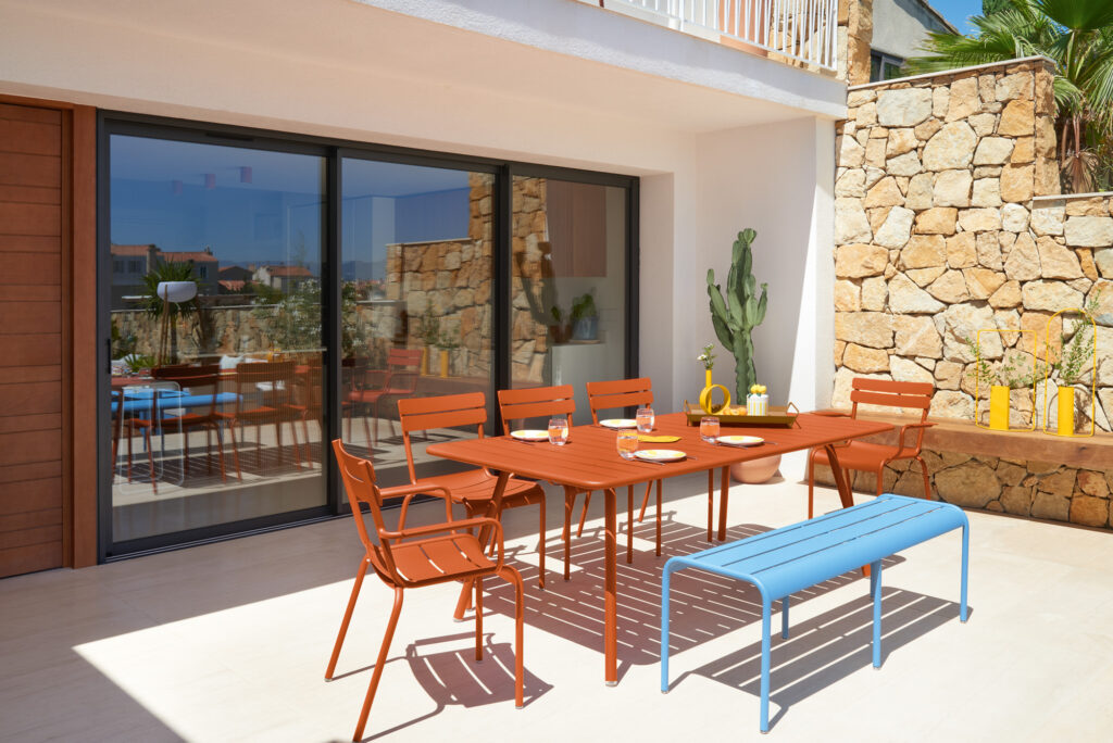 La terrasse est ornée d'une Table Luxembourg rouge vif de 207 x 100 cm, de chaises et d'un confortable banc bleu. Un mur de pierre de 100 cm et des cactus saisissants se dessinent en arrière-plan, tandis que des portes vitrées reflètent le paysage. La table est dressée avec des assiettes et des pichets pour une mise en scène parfaite.