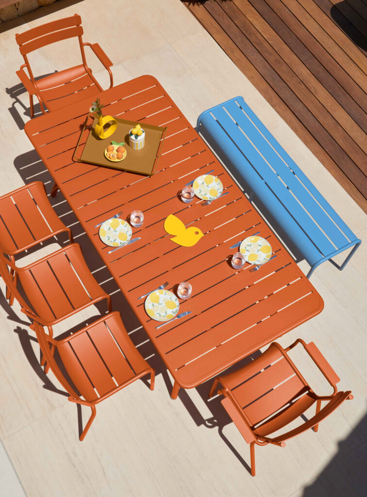 Une vue de dessus de la configuration de patio Table Luxembourg (207 x 100 cm) présente six chaises orange et un banc bleu, avec un plateau de boissons, des cupcakes, quatre assiettes et une décoration fantaisiste de canard en caoutchouc.