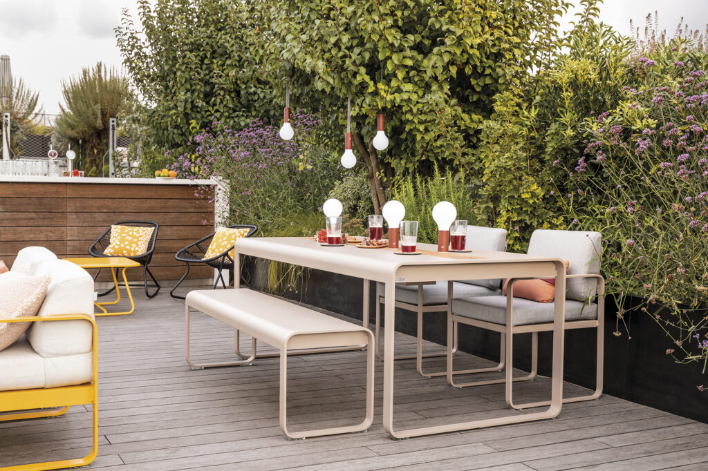 Un patio extérieur moderne met en valeur le banc Banc Bellevie avec une table et des chaises beiges, décorées de boissons et d'ampoules, entourées de plantes et de fleurs luxuriantes.