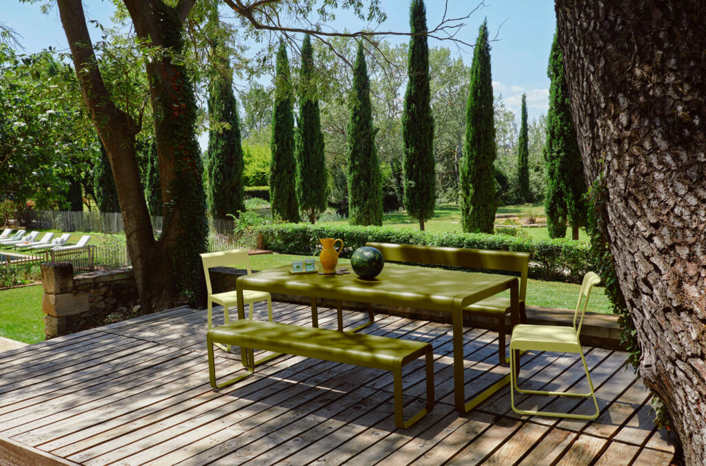 Ensemble de salle à manger d'extérieur en métal vert Banc Bellevie avec bancs et chaise sur une terrasse en bois, entourée d'arbres et donnant sur un jardin paysager avec de grands cyprès.