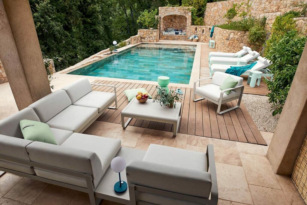 Élégant patio extérieur avec une Table Basse Bellevie de 103 x 75 cm donnant sur une piscine sereine entourée de verdure luxuriante et de murs en pierre.