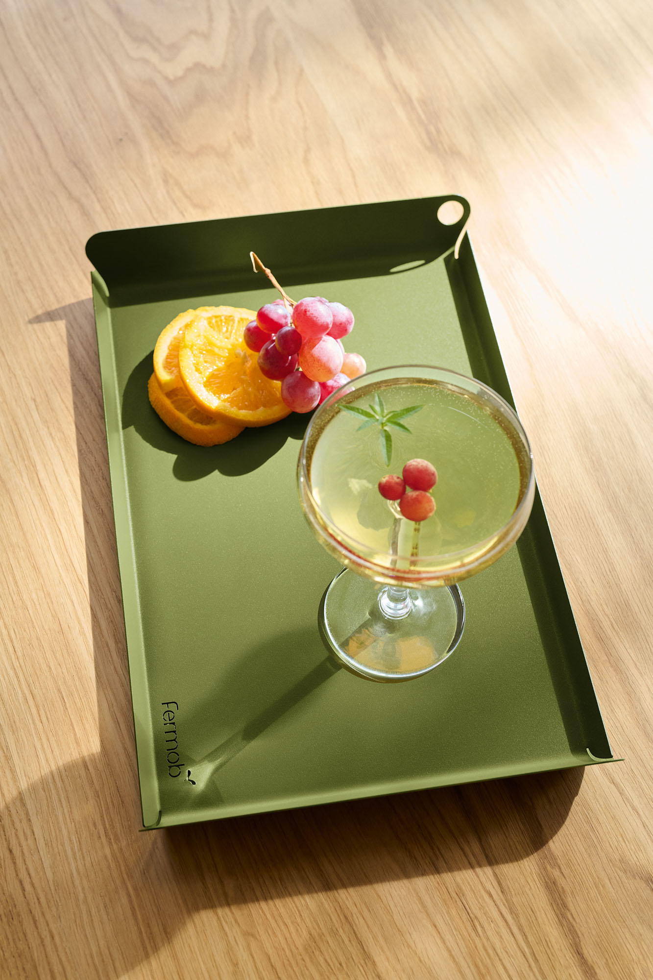 Un plateau Plateau Alto, 36x23 cm de couleur verte, contient un cocktail garni de raisins et de romarin, placé à côté de tranches d'orange sur une surface en bois clair.