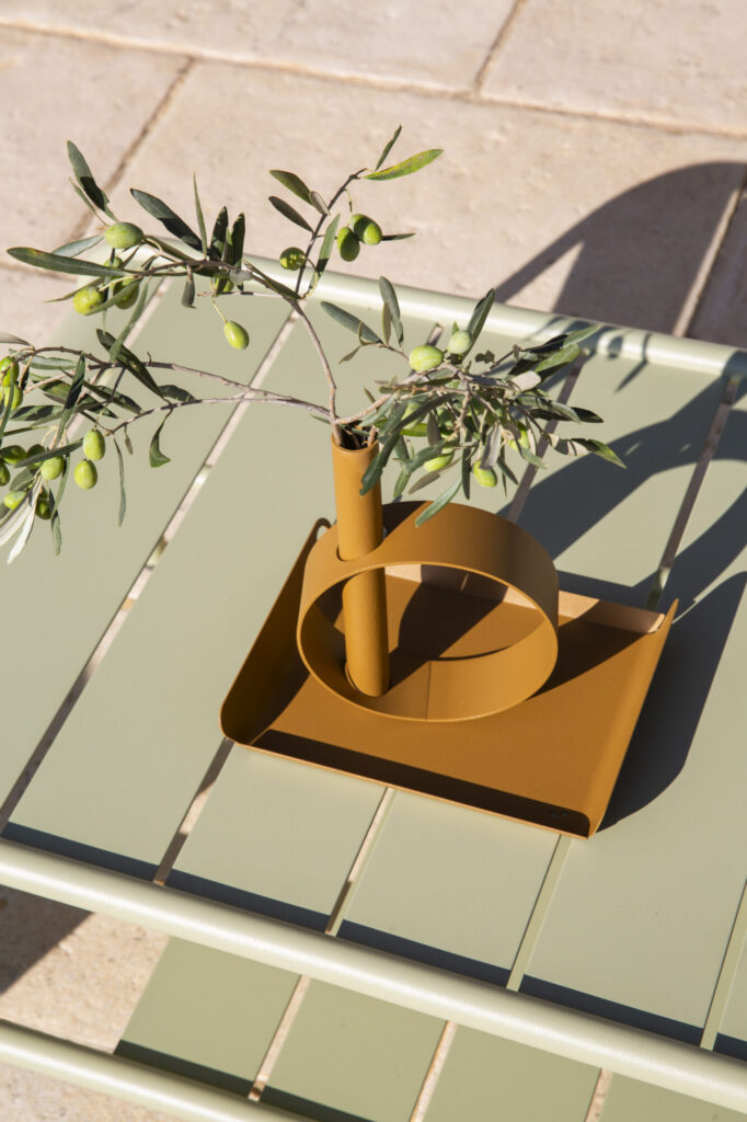 Une branche d'olives vertes est posée dans un vase moderne en métal jaune moutarde sur la table d'extérieur à lattes vert clair Plateau Alto 36x23 cm.