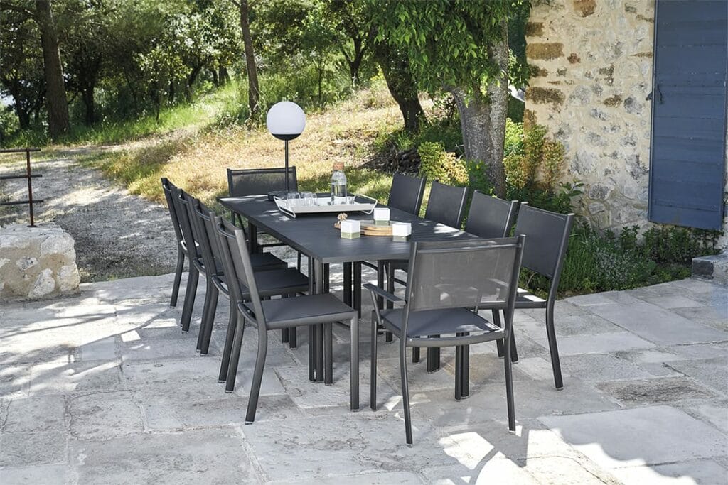 Coin repas extérieur avec une table et des chaises Costa extensibles modernes situées sur un patio en pierre, adjacent à un bâtiment en pierre avec une porte bleue, entouré d'arbres.