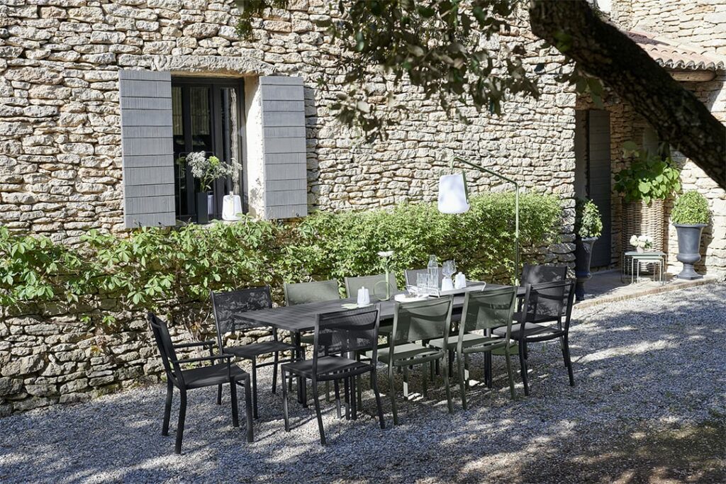 Salle à manger extérieure sous un arbre avec vue sur une maison en pierre recouverte de lierre, comprenant une table extensible Costa et des chaises sur un sol en gravier.