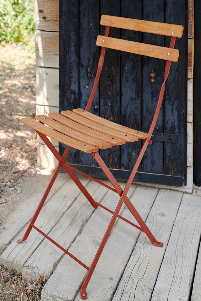 La Chaise Bistro naturel, une chaise pliante en bois de style bistrot avec des pieds en métal, trône gracieusement sur le porche en bois à côté d'une porte patinée.