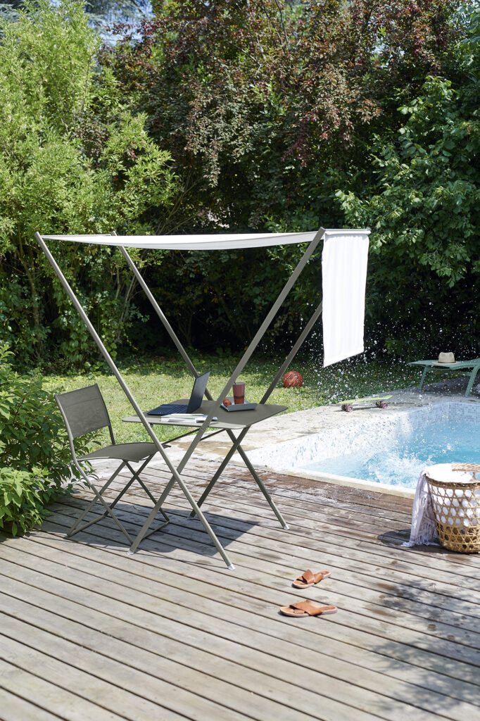 Deux chaises Chaise Plein Air et une petite table reposent sous un auvent sur une terrasse en bois au bord de la piscine, avec des sandales au sol et une serviette suspendue à proximité.