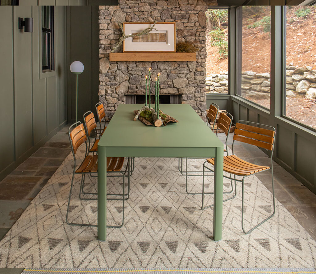 Une table extensible Ribambelle - 2 allonges en vert, associée à six chaises en bois, se dresse sur un tapis à motifs près d'une cheminée en pierre avec décor de manteau et de grandes fenêtres encadrant un paysage rocheux.