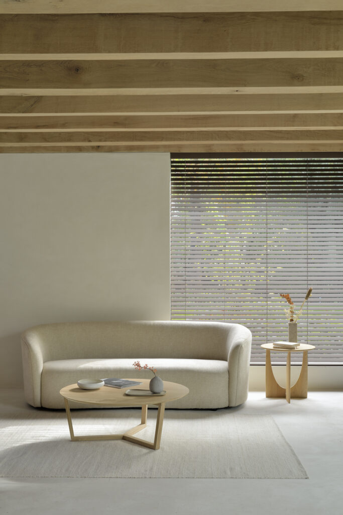 Un salon minimaliste met en valeur un canapé beige aux côtés d'une table basse Tripod en chêne chic. Une table d'appoint avec des vases renforce l'élégance, tandis que la grande fenêtre avec stores invite la lumière naturelle. Des poutres au plafond et un tapis clair complètent le décor, créant une atmosphère sereine.