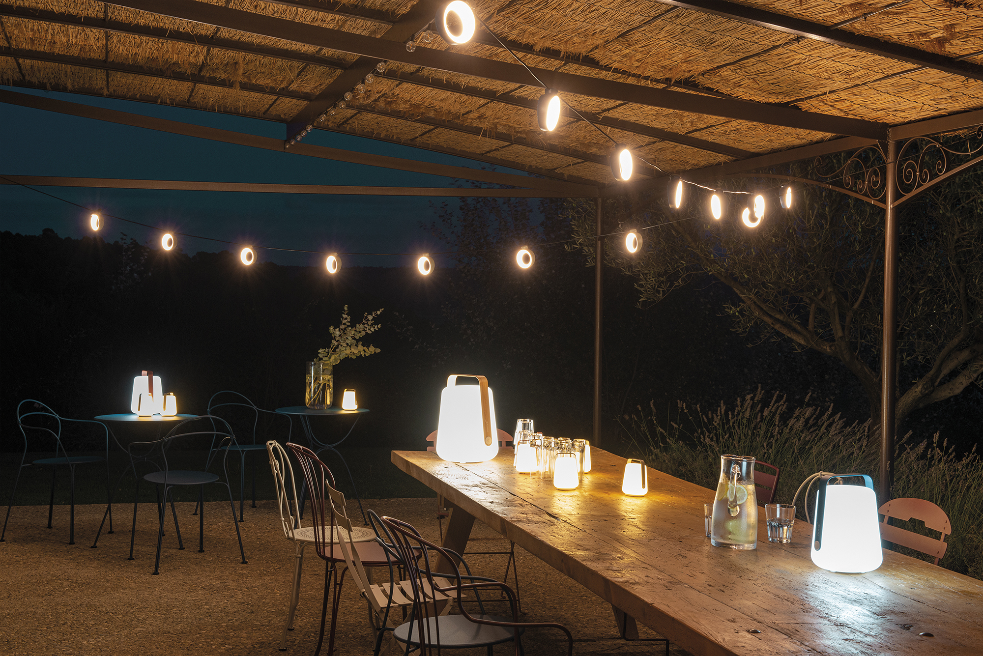 La nuit, le patio extérieur dispose d'une table en bois avec des pichets à boissons, des verres et des lanternes portables ; les guirlandes lumineuses Guirlande Hoop brillent au-dessus tandis que plusieurs chaises entourent des tables plus petites à proximité.