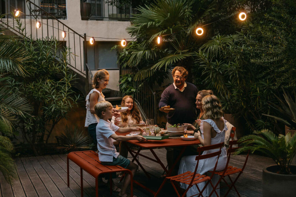 Six personnes, dont deux enfants, se rassemblent autour d'une table extérieure dressée pour le dîner au crépuscule, entourées de plantes et illuminées par les guirlandes lumineuses Guirlande Hoop drapées au-dessus.