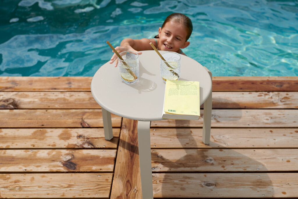 Une petite fille debout sur une terrasse en bois au bord d’une piscine, tenant une table basse Alize.