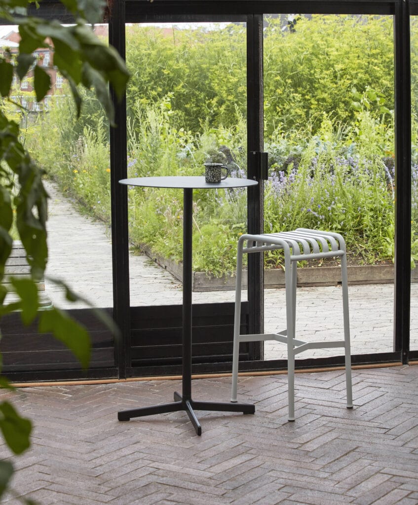 Une table avec une plante dessus devant une fenêtre. Le Tabouret haut Palissade est placé à côté de la fenêtre, créant un coin cosy et invitant. La Palissade sert de