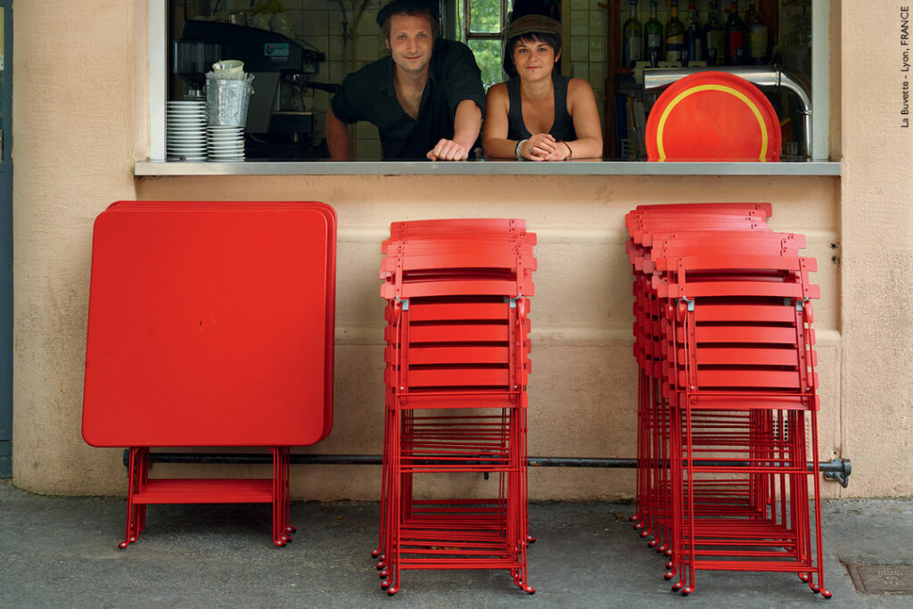 Deux personnes se tiennent derrière la vitrine d’un café. Des piles de chaises rouges et une Table carrée Bistro pliée, 71 x 71 cm, sont placées à l'extérieur.