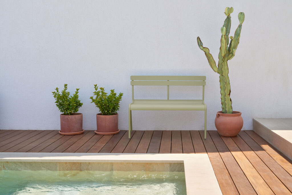 Une scène de patio minimaliste présente un Banc vert avec dossier Luxembourg, deux plantes en pot et un cactus sur une terrasse en bois près d'une petite piscine contre un mur blanc.
