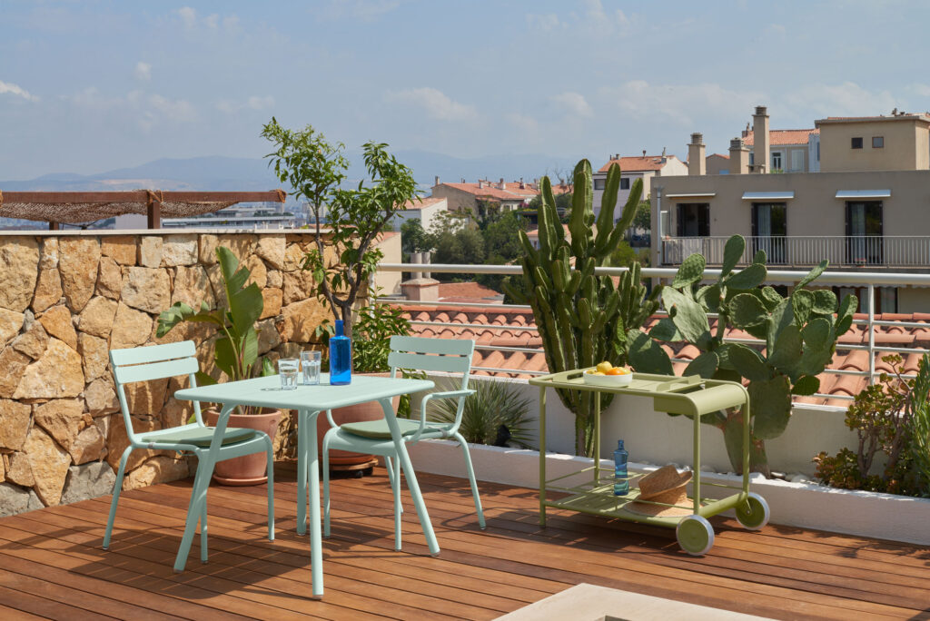 Sur le toit-terrasse, avec sa terrasse en bois, se trouve la Table carrée Luxembourg, d'un mètre vingt pour deux personnes, ainsi qu'une plante en pot, un cactus et un chariot à roulettes. Le ciel luxembourgeois partiellement nuageux est en toile de fond, avec des bâtiments en arrière-plan.