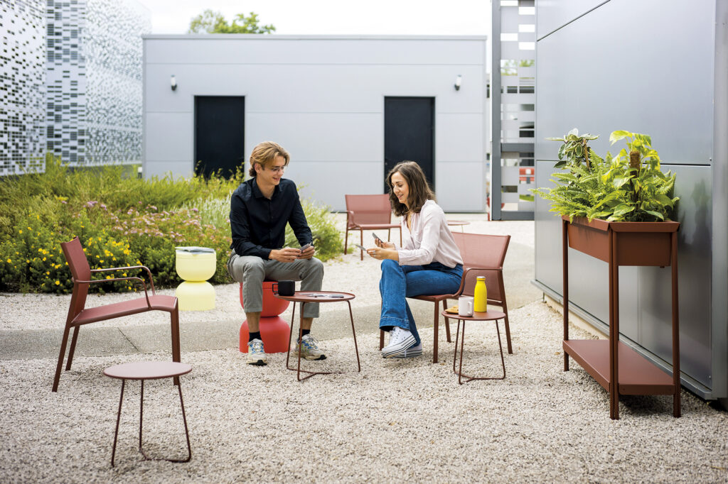Deux personnes discutent et jouent aux cartes à une petite table extérieure sur une terrasse en gravier, entourées de chaises modernes, de plantes luxuriantes dans la Grande jardinière Terrazza et d'une bouteille sur la table.