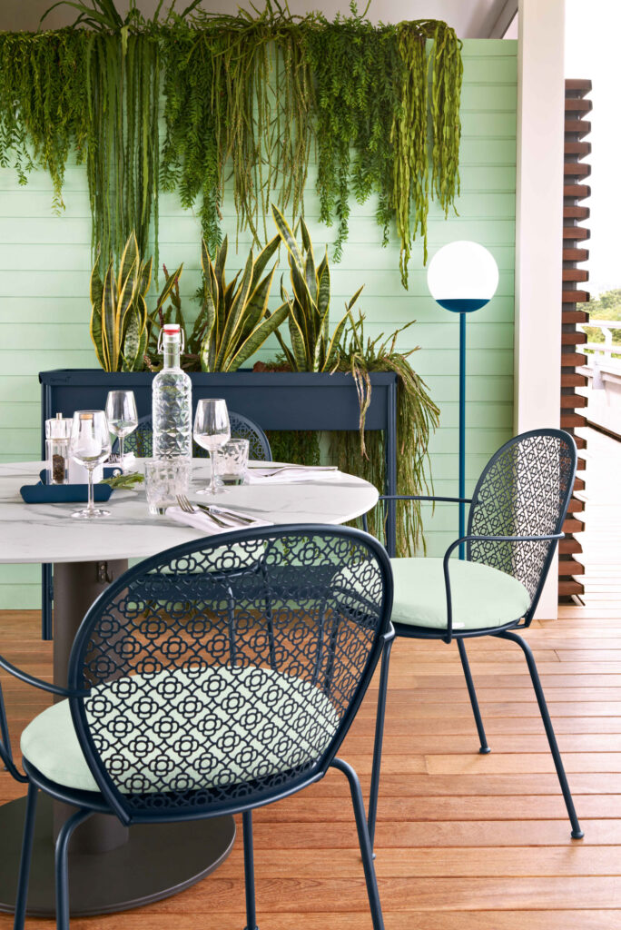 Un espace repas extérieur moderne avec une table ronde, des chaises en métal à motifs, de la verrerie, une bouteille et un mur vert luxuriant orné de plantes suspendues et de la Grande jardinière Terrazza en arrière-plan.