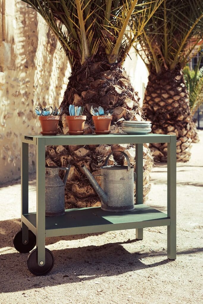 Une Desserte Quiberon avec des plantes grasses en pot et des arrosoirs sous un palmier, dans un cadre extérieur ensoleillé.