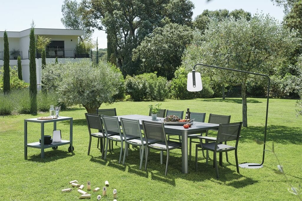 Coin repas extérieur moderne avec une grande table, des chaises et une Desserte Quiberon suspendue dans un jardin luxuriant à côté d'une maison de style contemporain.
