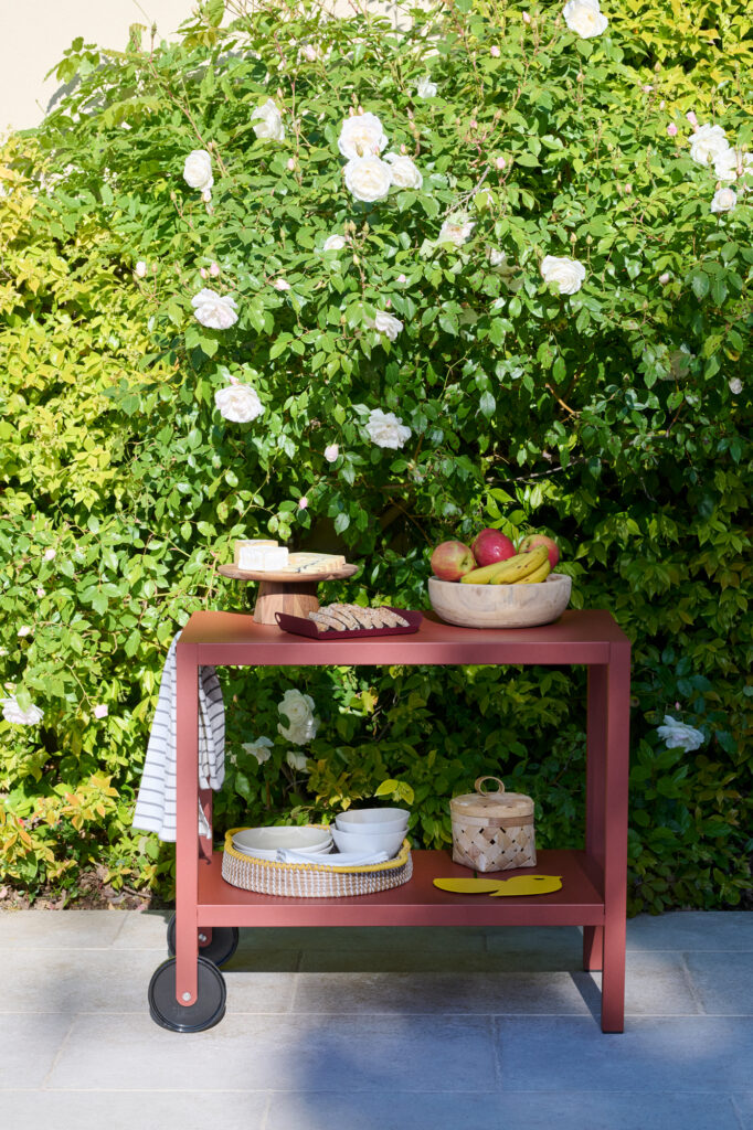Un chariot rouge Desserte Quiberon contenant des assiettes, des bols, une coupe de fruits et divers articles de table est exposé à l'extérieur devant un buisson vert luxuriant parsemé de fleurs blanches.