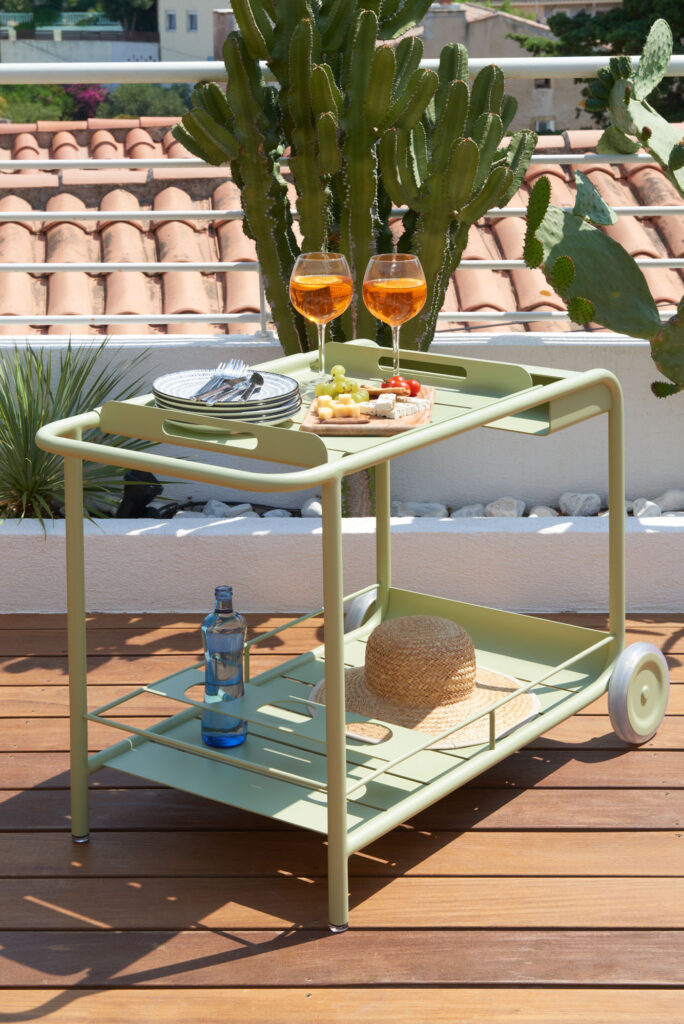 L'élégant Desserte Luxembourg orne la terrasse en bois, proposant boissons, assiettes et en-cas. Un cactus en arrière-plan s'harmonise avec les tuiles en terre cuite du toit, créant une ambiance chaleureuse.