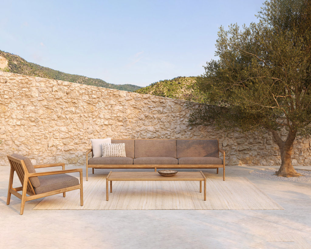 Un ensemble de patio Canapé Jack en bois de teck, comprenant un canapé 3 places et des chaises en bois, adossé à un mur de pierre et ombragé par un arbre.