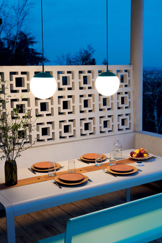 Une table Bellevie moderne avec rangement (196 x 90 cm) accueille quatre personnes avec assiettes, verres et couverts sous deux suspensions sphériques au crépuscule. Un vase à branches et une coupe à fruits apportent charme et rangement à la pièce.