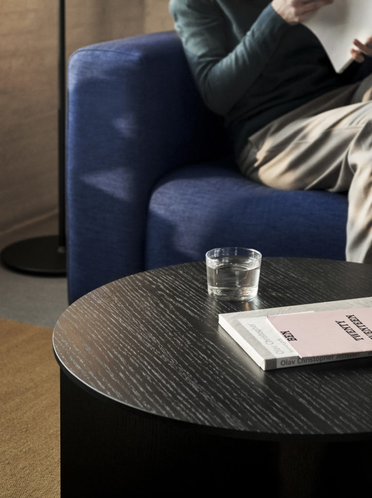 Un homme assis sur un canapé lisant un livre sur une Table d’appoint ronde en bois fendu.