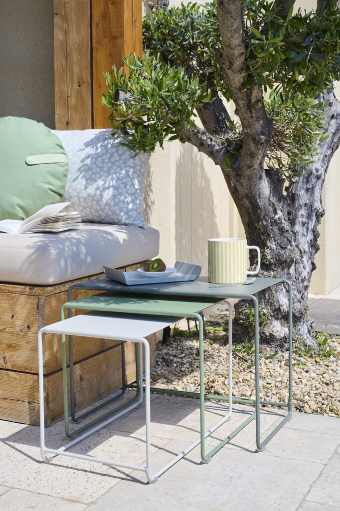 Set de 3 tables basses Oulala avec une tasse rayée, une assiette et une cuillère, près d'un banc en bois et d'un olivier sur un patio ensoleillé.