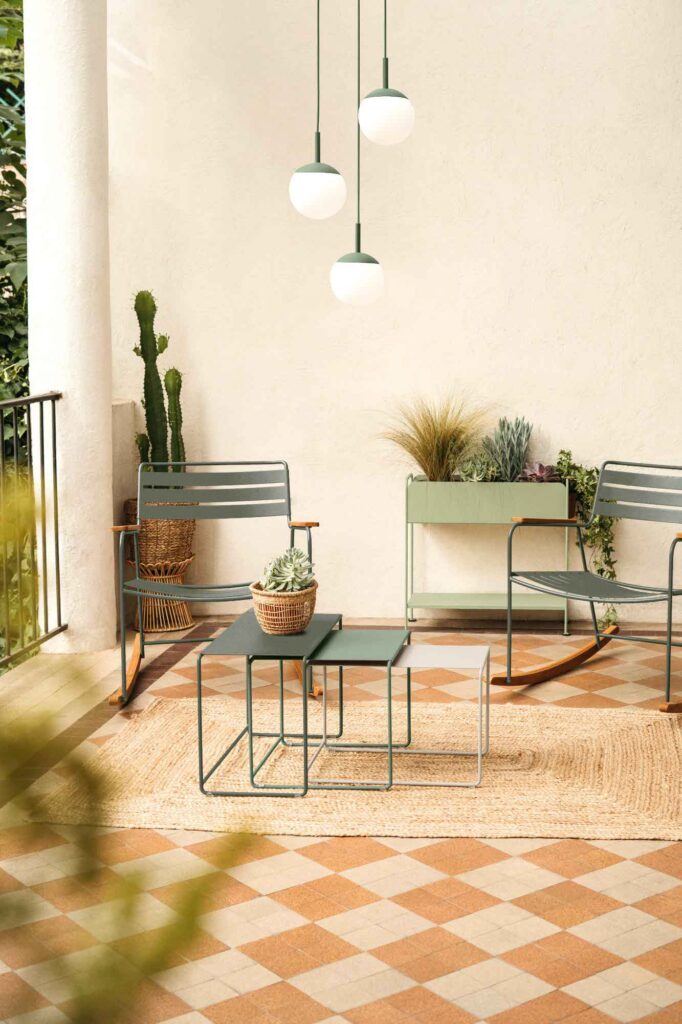 Patio extérieur avec deux chaises en métal vert, un set de 3 tables basses Oulala, des plantes en pot et trois lampes suspendues contre un mur de couleur claire.