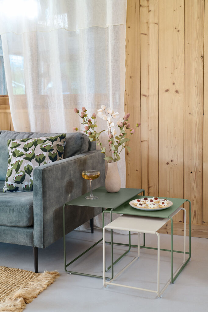 Un salon moderne est composé d'un canapé gris, d'un coussin à motifs et d'un Set de 3 tables basses Oulala en vert tenant un vase de fleurs, une boisson et des amuse-gueules, sur fond de rideaux clairs et de murs en bois.