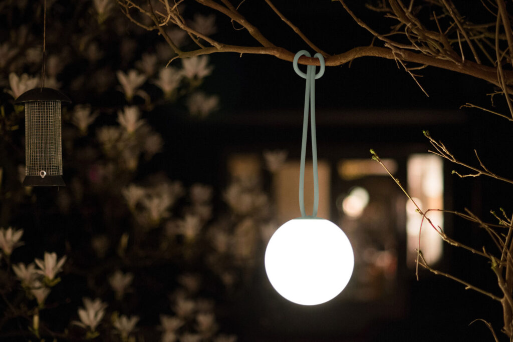 Une lanterne ronde est suspendue à un arbre avec une sangle bleue près d'une mangeoire à oiseaux ; en dessous repose le Fauteuil pour salon de jardin Paletti - 1 place, entouré de fleurs et de fenêtres floues à l'arrière-plan.