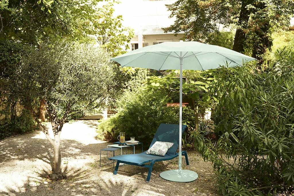 Un jardin tranquille comprenant une chaise longue bleue sous un parasol Shadoo bleu clair, entouré d'une verdure luxuriante et d'arbres, avec une petite table contenant des livres et une tasse.