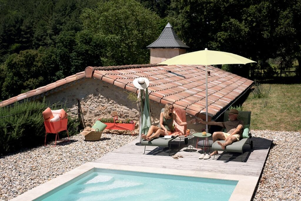 Un couple se détend sur des chaises longues au bord d'une petite piscine, avec un parasol Shadoo et une verdure pittoresque en arrière-plan.