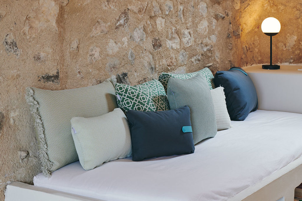 Un Coussin Color Mix 44x30cm coloré sur un lit dans une chambre avec un mur en pierre.