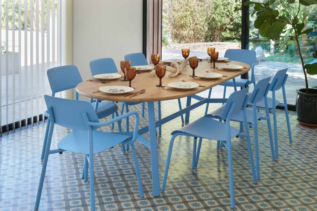 Une table à manger dressée pour huit personnes avec des verres et des assiettes ambrés, entourée de chaises bleues Chaise Studie dans une pièce lumineuse avec des sols carrelés à motifs et de grandes fenêtres donnant sur un jardin.