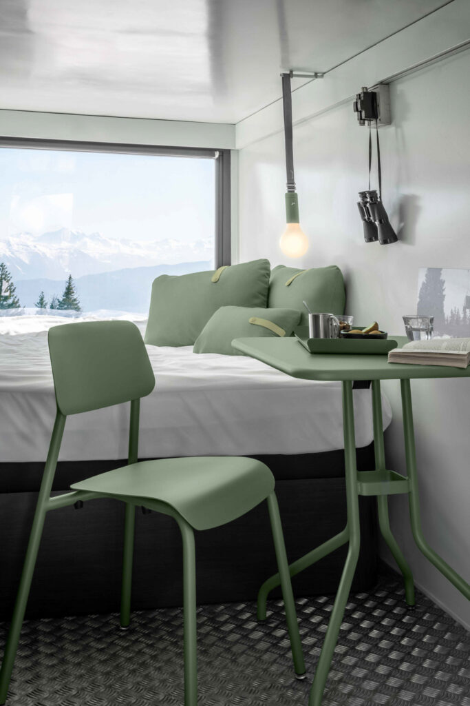 Une petite chambre minimaliste avec une chaise longue verte, une table assortie, un lit avec des oreillers verts, une ampoule suspendue et une fenêtre présentant des montagnes enneigées et des arbres à l'extérieur.