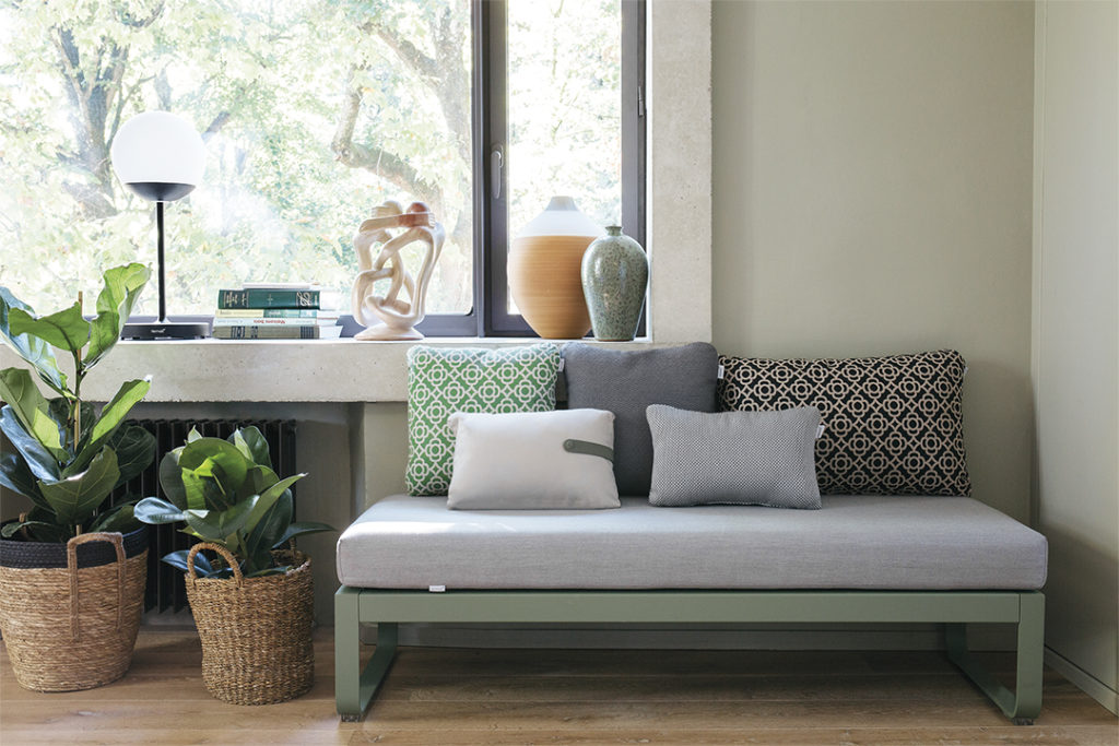 Un canapé avec des coussins et des plantes devant une fenêtre, éclairé par la douce lueur de la Lampe Mooon ! M.