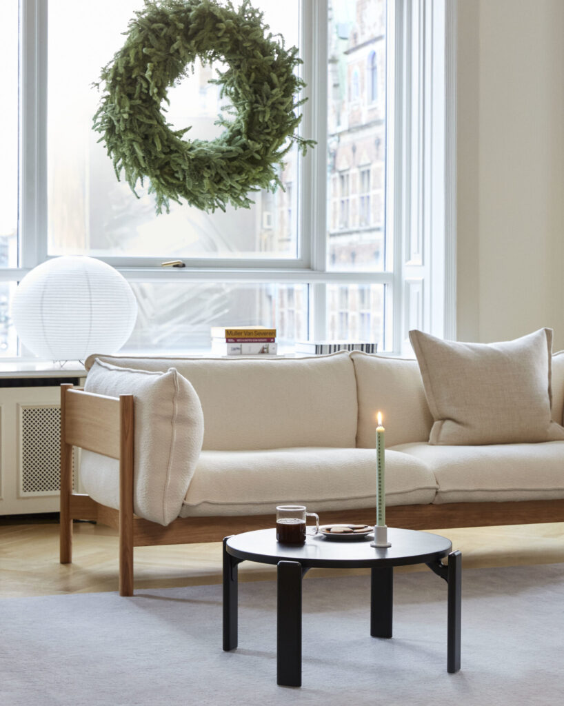 Un salon confortable comprend un canapé beige, un tapis bleu ardoise sous une table basse ronde noire avec une bougie allumée et une boisson, et une grande couronne verte suspendue devant la fenêtre.