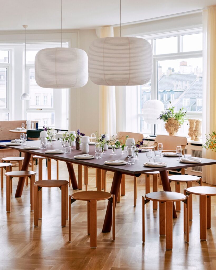 Une salle à manger spacieuse avec une longue table en bois dressée pour un repas, entourée de chaises Tabouret Rey simples. De grandes lanternes en papier pendent au-dessus, projetant une lueur chaleureuse, tandis que des touches de rouge écarlate ajoutent du style. De grandes fenêtres offrent une vue sur les bâtiments extérieurs.