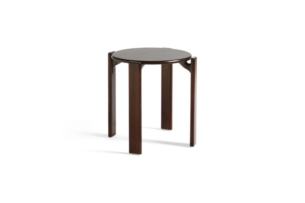 Un Tabouret Rey rond, marron foncé, doté de quatre pieds droits, se dresse élégamment sur un fond blanc, son design subtil offrant un contraste parfait avec les accents rouge écarlate à proximité.