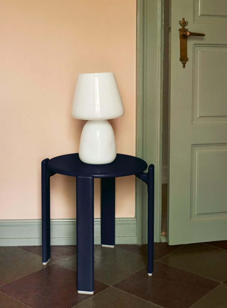 Une lampe blanche est posée sur un Tabouret Rey, une petite table ronde noire, dans le coin d'une pièce au mur rouge écarlate et à la porte gris clair.