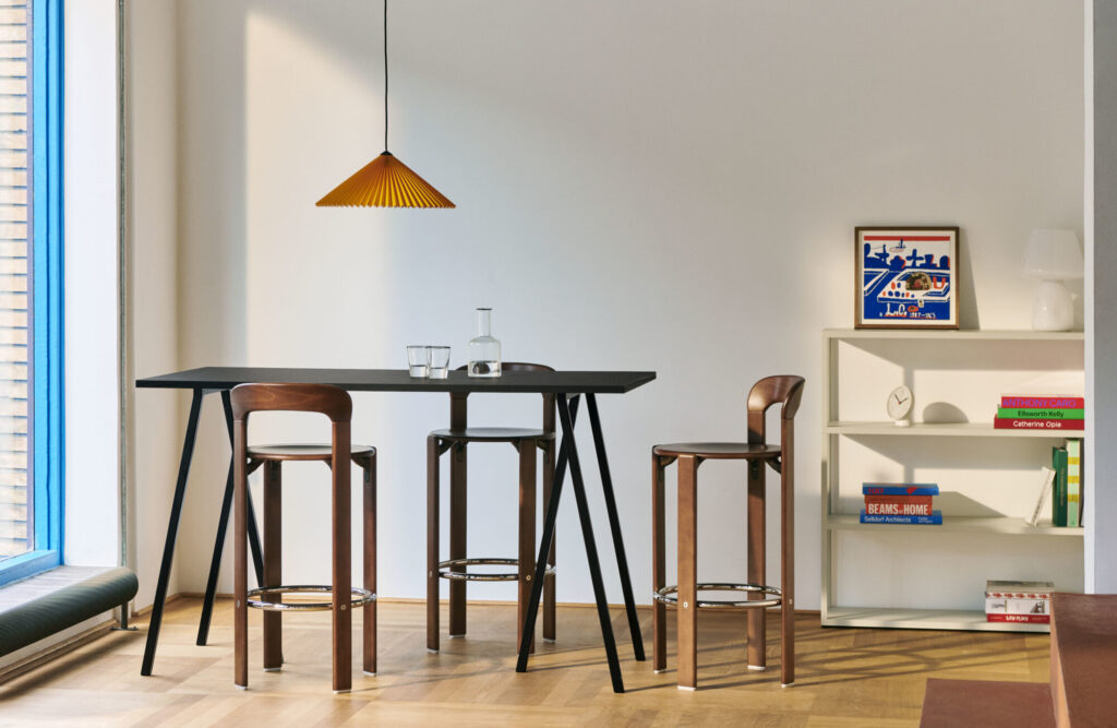Une salle à manger moderne est dotée d'une grande table noire accompagnée de trois tabourets hauts en bois, tandis qu'une lampe suspendue rouge écarlate est suspendue au-dessus. Contre le mur blanc se trouve une étagère ornée de livres et de décorations, mêlant harmonieusement style et fonctionnalité.