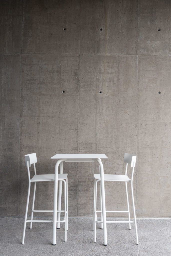 Une table de bar blanche August et des chaises devant un mur de béton dans un bar.