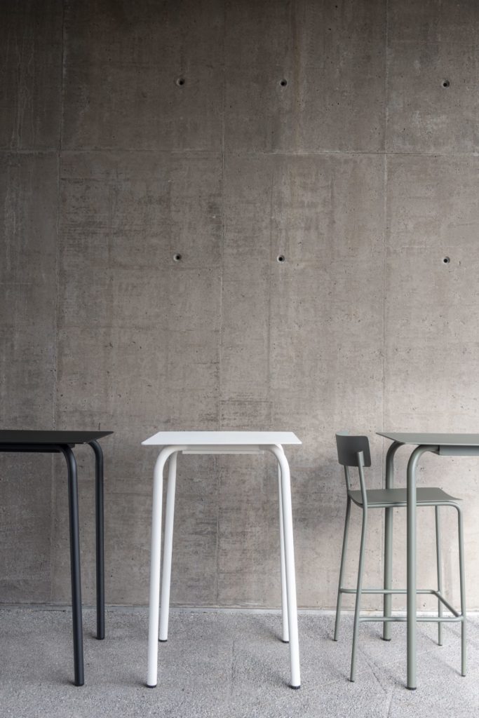 Une Table de bar August et des tabourets installés devant un mur de béton.