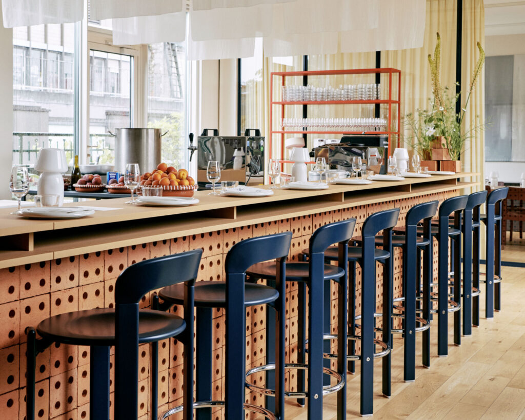 Un bar moderne avec des tabourets bleus Tabouret haut Rey, un comptoir en bois avec des assiettes, des verres, des coupes de fruits, des étagères de verrerie et des vues sur la ville à travers de grandes fenêtres.