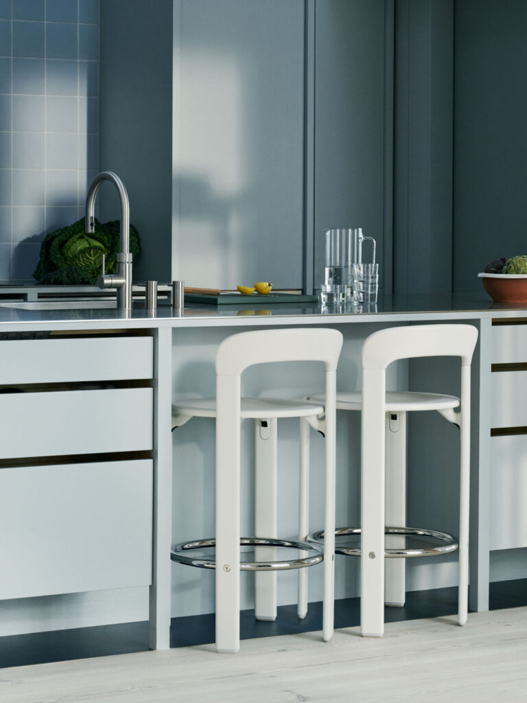 Une cuisine moderne avec des armoires bleu clair, deux tabourets Tabouret haut Rey blancs sous l'îlot, un évier avec robinet, des verres sur le comptoir et des légumes frais en arrière-plan.