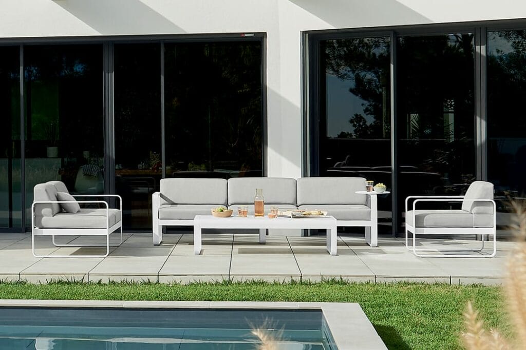 Canapé d'extérieur moderne 3 places Bellevie au bord d'une piscine, composé d'un canapé et de deux chaises aux coussins blancs, d'une table basse avec des collations, sur fond de maison aux parois de verre.