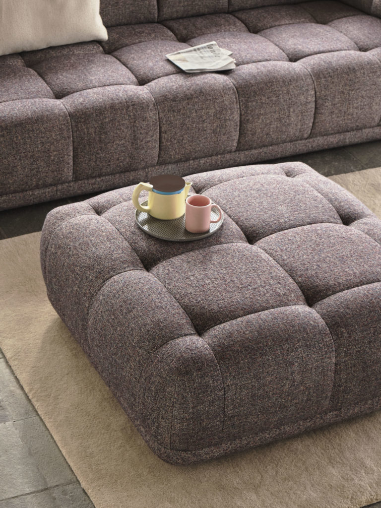 Pouf Quilton ottoman de HAY en tissu ou cuir de la marque Hay dans un salon sur un tapis avec une assiette et tasses sur le pouf